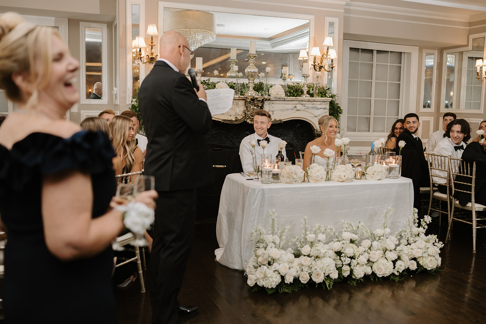 Newlyweds laugh from their white floral sweetheart table as the bride's father delivers a heartwarming toast.